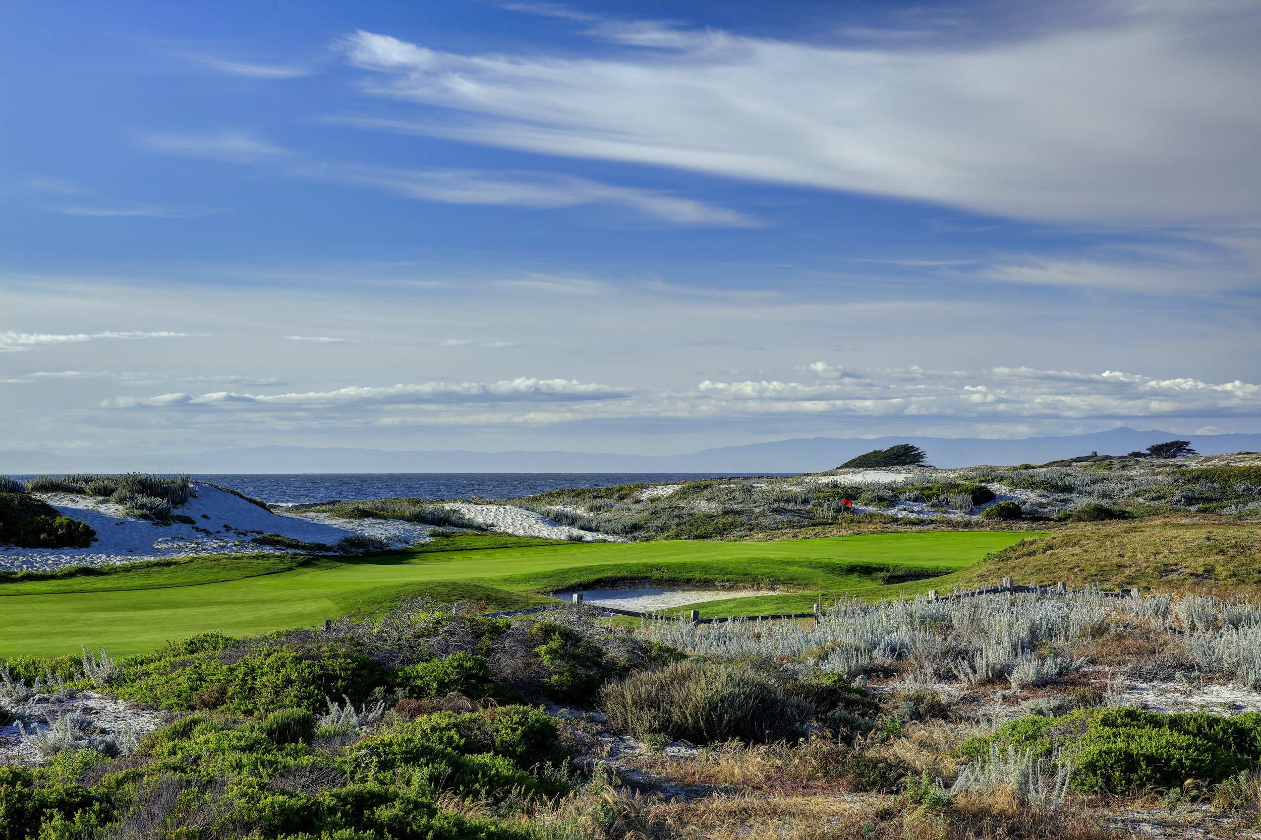 A green golf course surrounded with short shrubs and the ocean in the distance on a partly cloudy day.