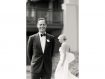 Black and white photo of groom in tux with bride in background