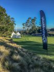 Golf green with two white tents, a large group of people and trees in the background on a sunny day.