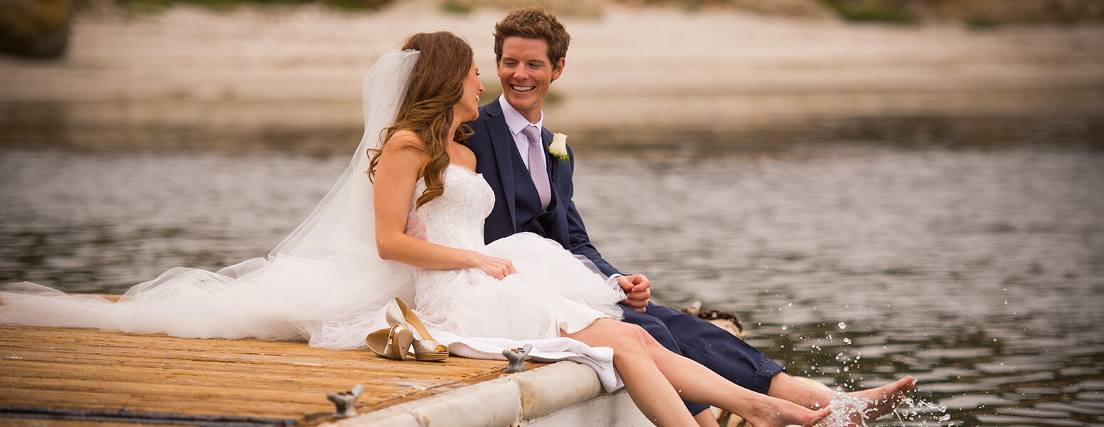 Bride and a groom sitting on a dock with their feet in the water.